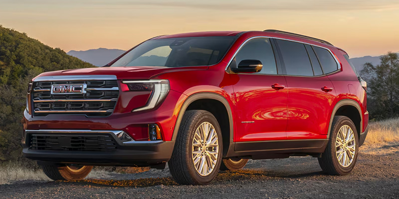 Passenger side view of a red 2025 GMC Acadia parked with the sun setting behind