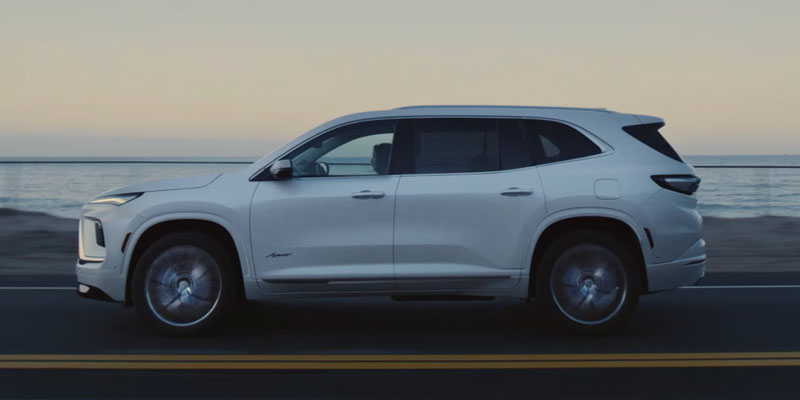 White 2026 Buick Enclave driving along a coastal highway at sunset with ocean views in the background.