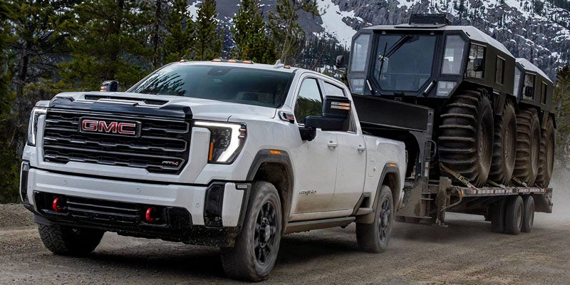 2026 GMC Sierra towing a heavy-duty vehicle on a rugged dirt road with snow-capped mountains in the background.